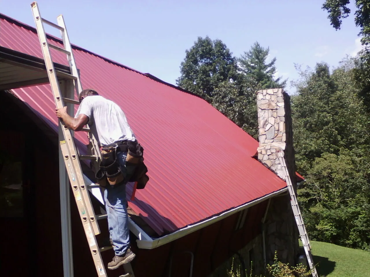 Roofer on a ladder beside a red metal roof and stone chimney — Soffit & Fascia Repair by Roofing Brothers in Cuthbert